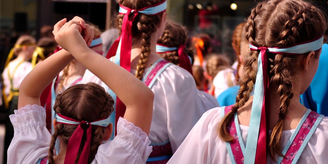 Kolme nuorta lettipäistä tyttöä kuvattu takaa päin perinnepuvut yllään..